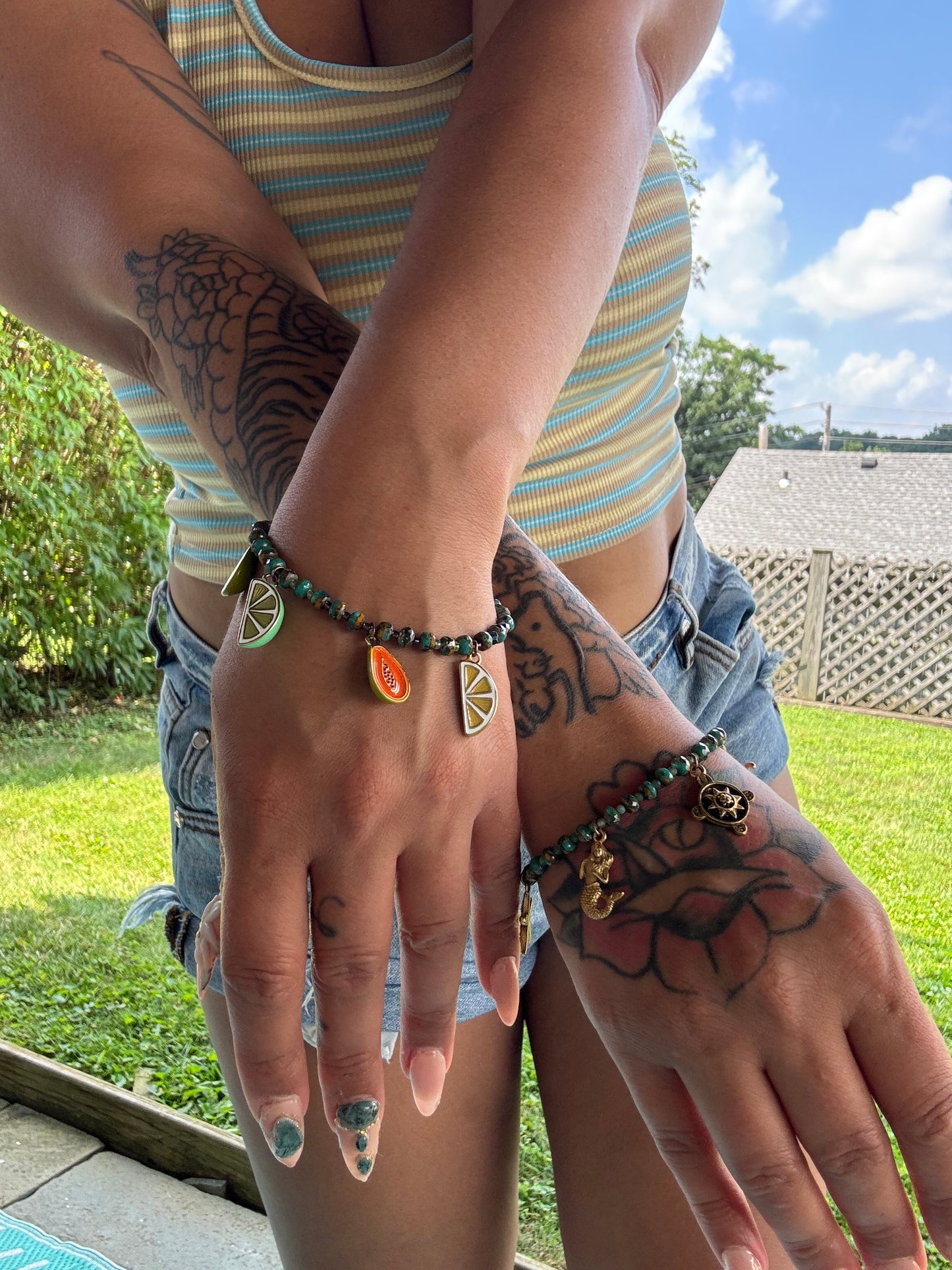 A close-up lifestyle shot of a person's tattooed arms and hands wearing two beaded bracelets outdoors in a backyard. One bracelet features green, orange, and yellow citrus slice charms, and the other is dark green with a small gold charm.