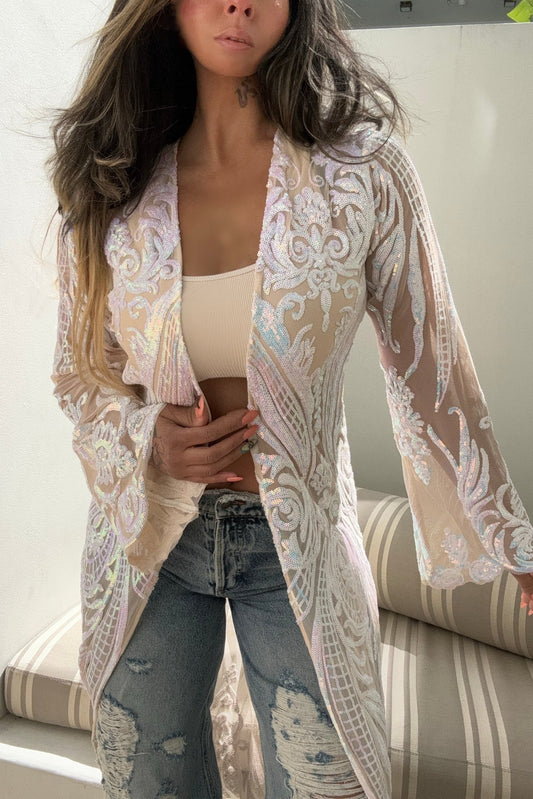 A woman wearing a long, sheer beige kimono-style cardigan with intricate white floral embroidery and iridescent pastel accents, paired with a beige tank top and distressed blue jeans.