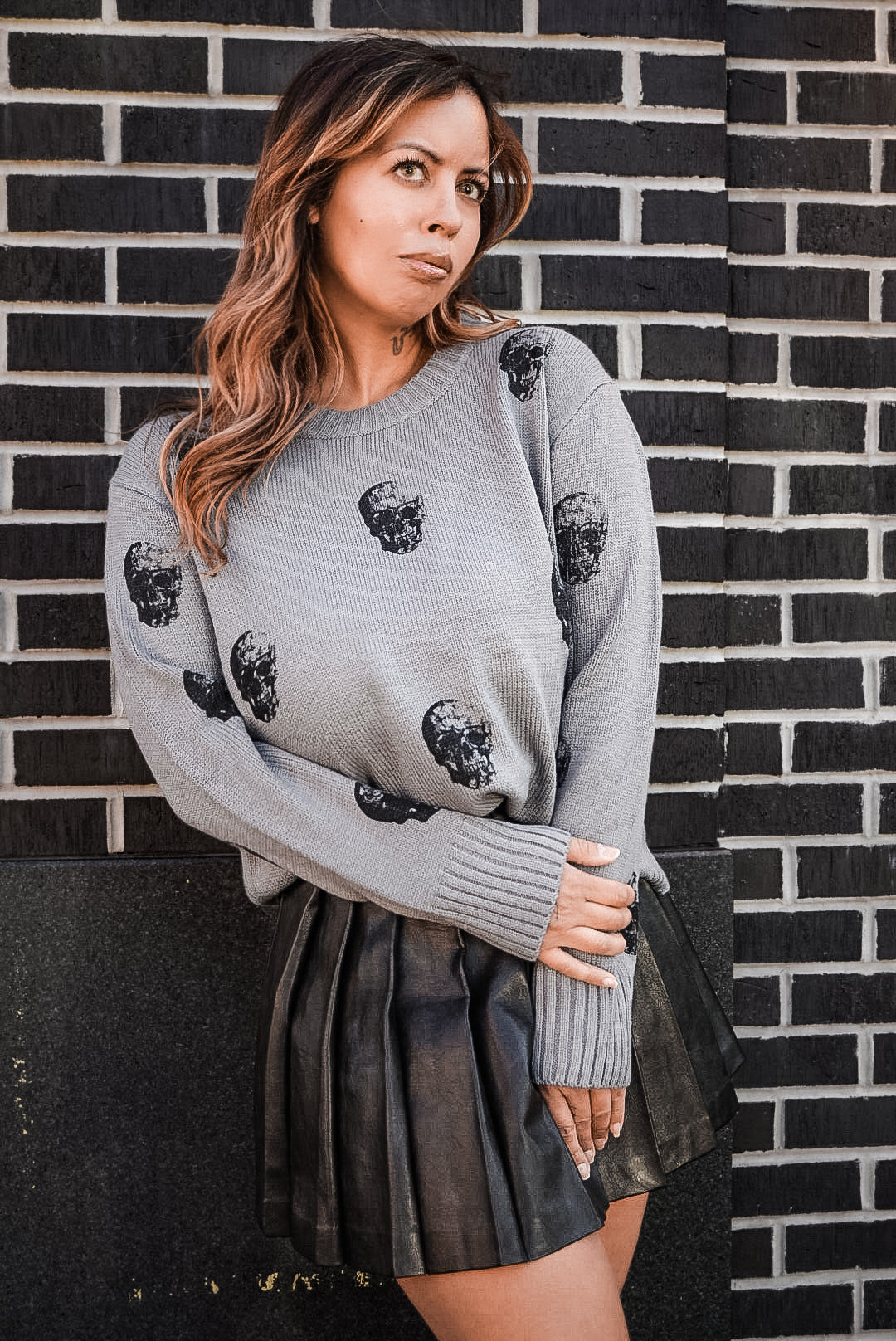 A woman wearing a light gray crewneck sweater with black skull prints and a dark pleated skirt, posing against a black brick wall.