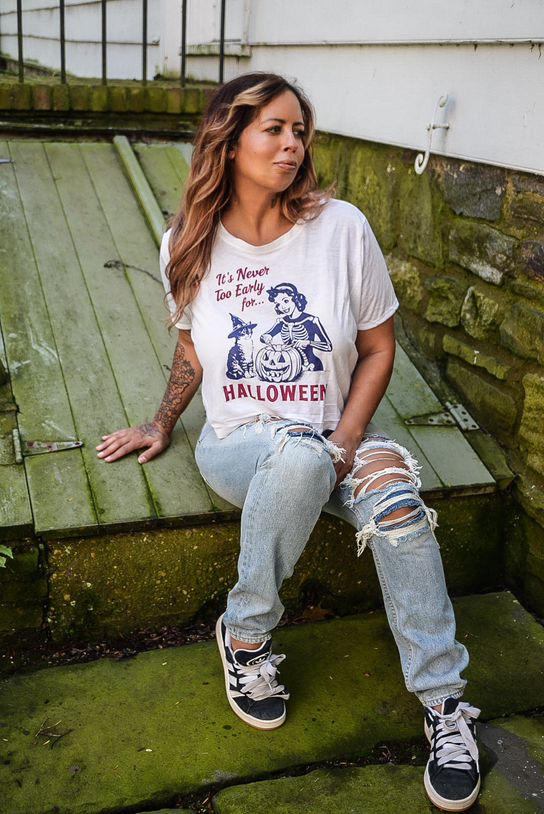 A woman sitting on mossy outdoor steps wearing a white graphic t-shirt with a Halloween design, distressed light blue jeans, and black sneakers.