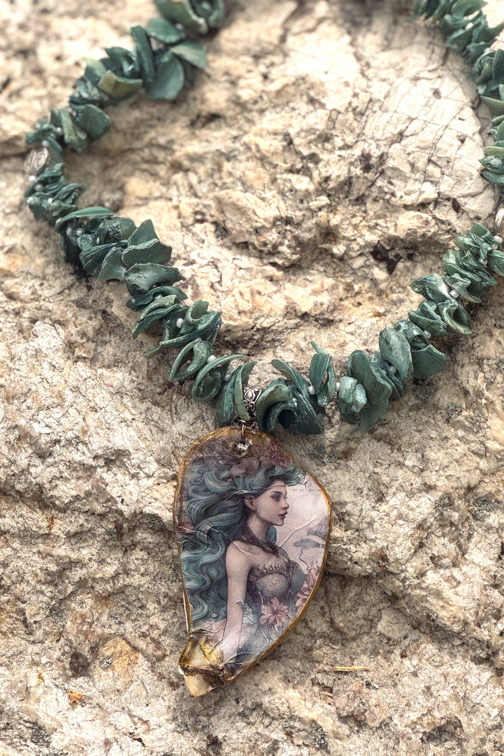 A necklace made of dark green chip-style beads with a teardrop pendant featuring a painted mermaid illustration, displayed on a textured beige rock surface.