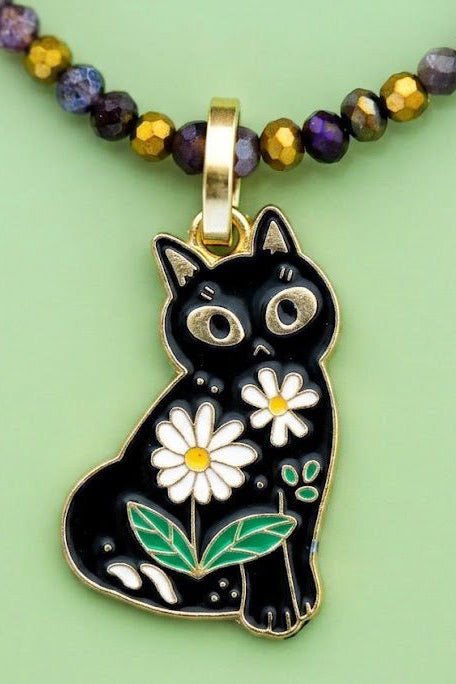 A black cat pendant necklace with white daisy flowers and green leaves, hanging from a beaded chain with purple, gold, and brown beads on a light green background.