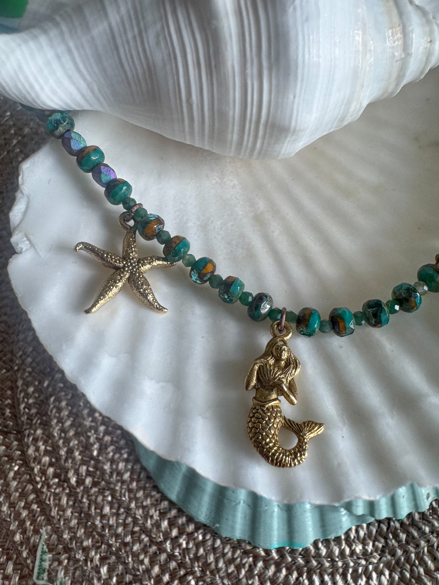 A beaded necklace with turquoise and iridescent beads, featuring a gold starfish charm and a gold mermaid tail charm, displayed on a white seashell.