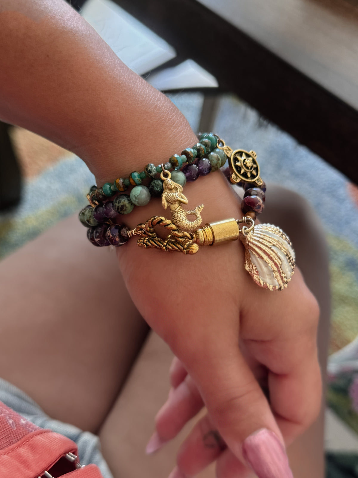 A close-up of a person's wrist wearing three beaded bracelets with gold-toned charms, including a mermaid, seashell, star, and a green stone bead strand.