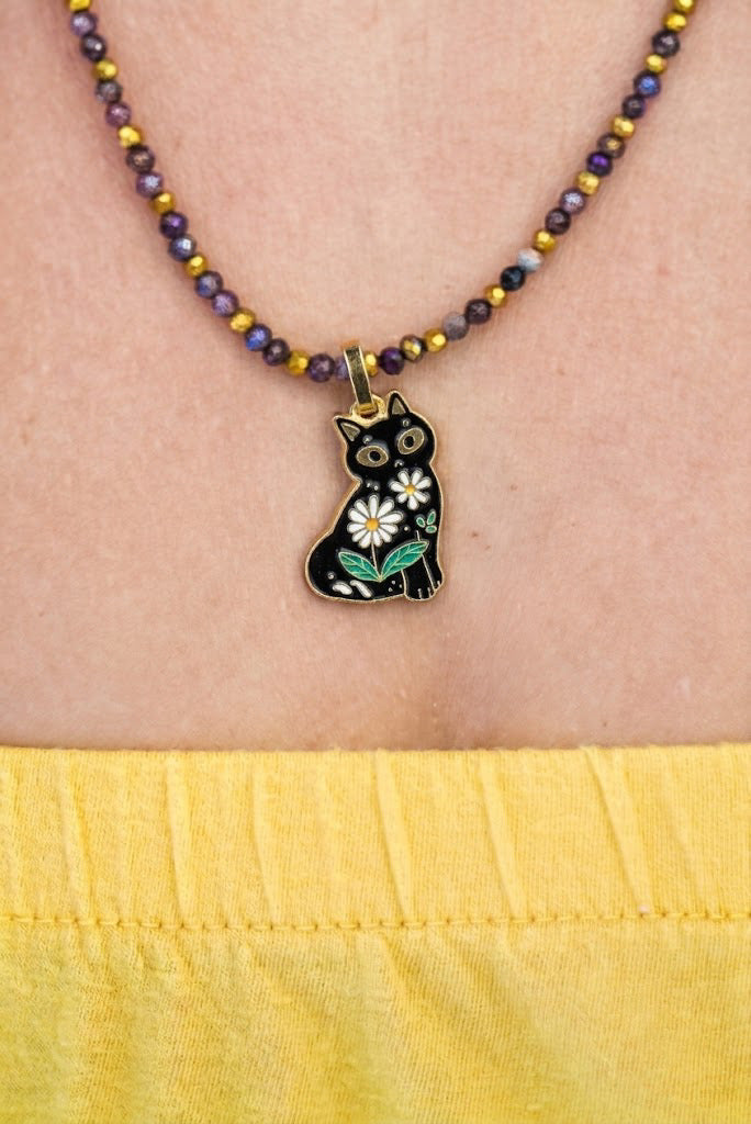 A close-up of a person wearing a beaded necklace with a black cat pendant decorated with white and green flowers. The necklace features purple and gold beads, and the person is wearing a yellow top.