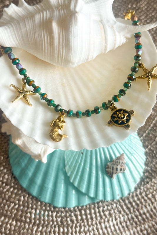A beaded necklace with green and purple beads, gold starfish charms, a mermaid charm, and a turtle charm, displayed on white and turquoise seashells.