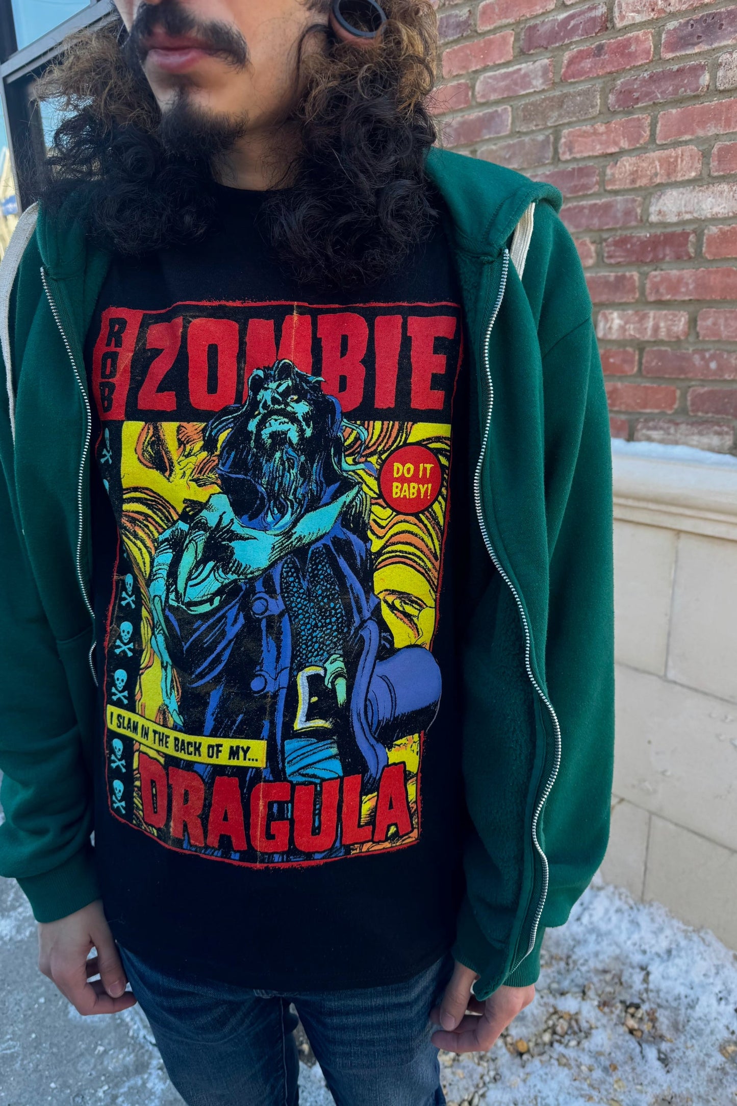A man wearing a black t-shirt with a graphic of a character and the text 'ZOMBIE DRAGULA' standing in front of a brick wall.