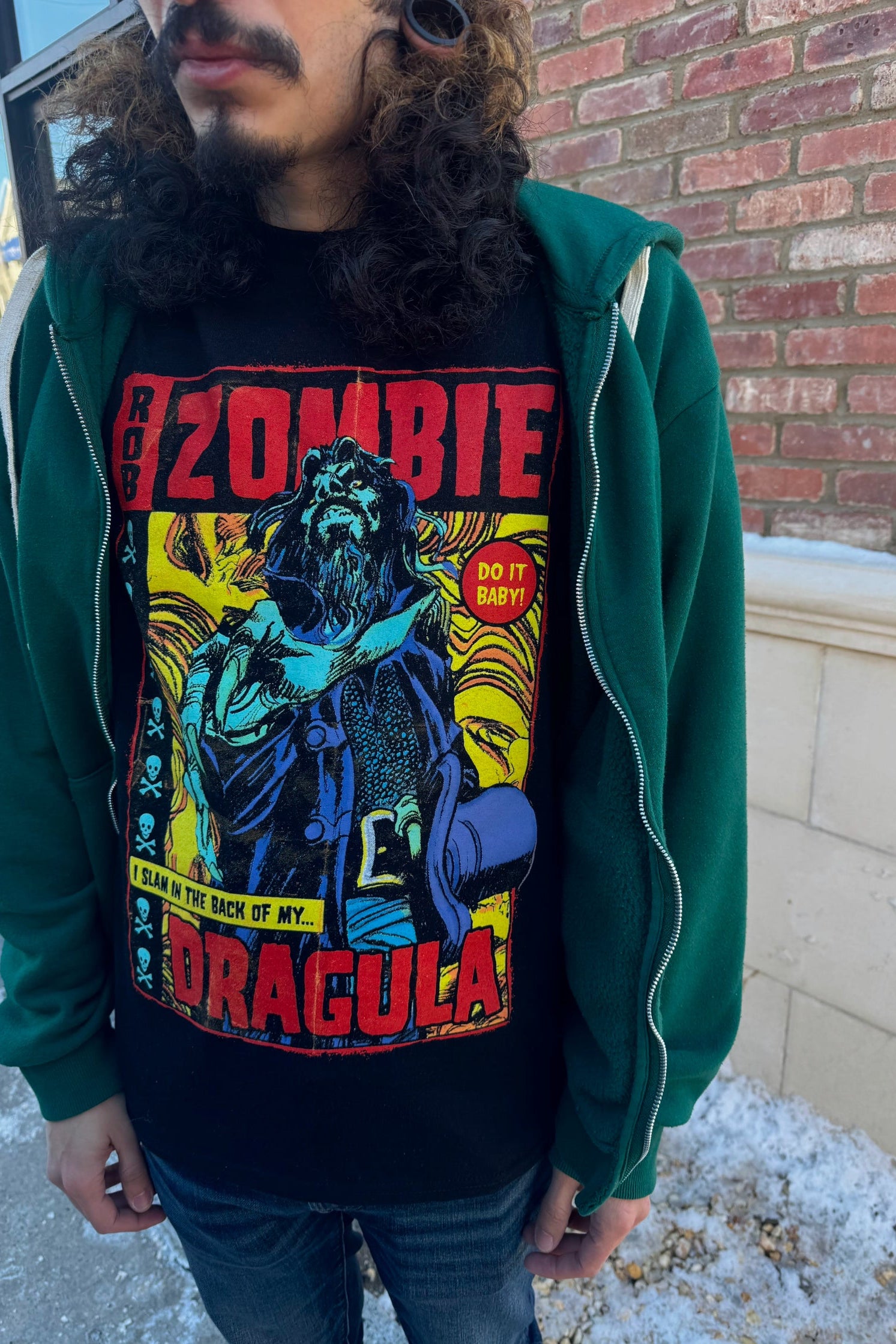 A man wearing a black t-shirt with a graphic of a character and the text 'ZOMBIE DRAGULA' standing in front of a brick wall.