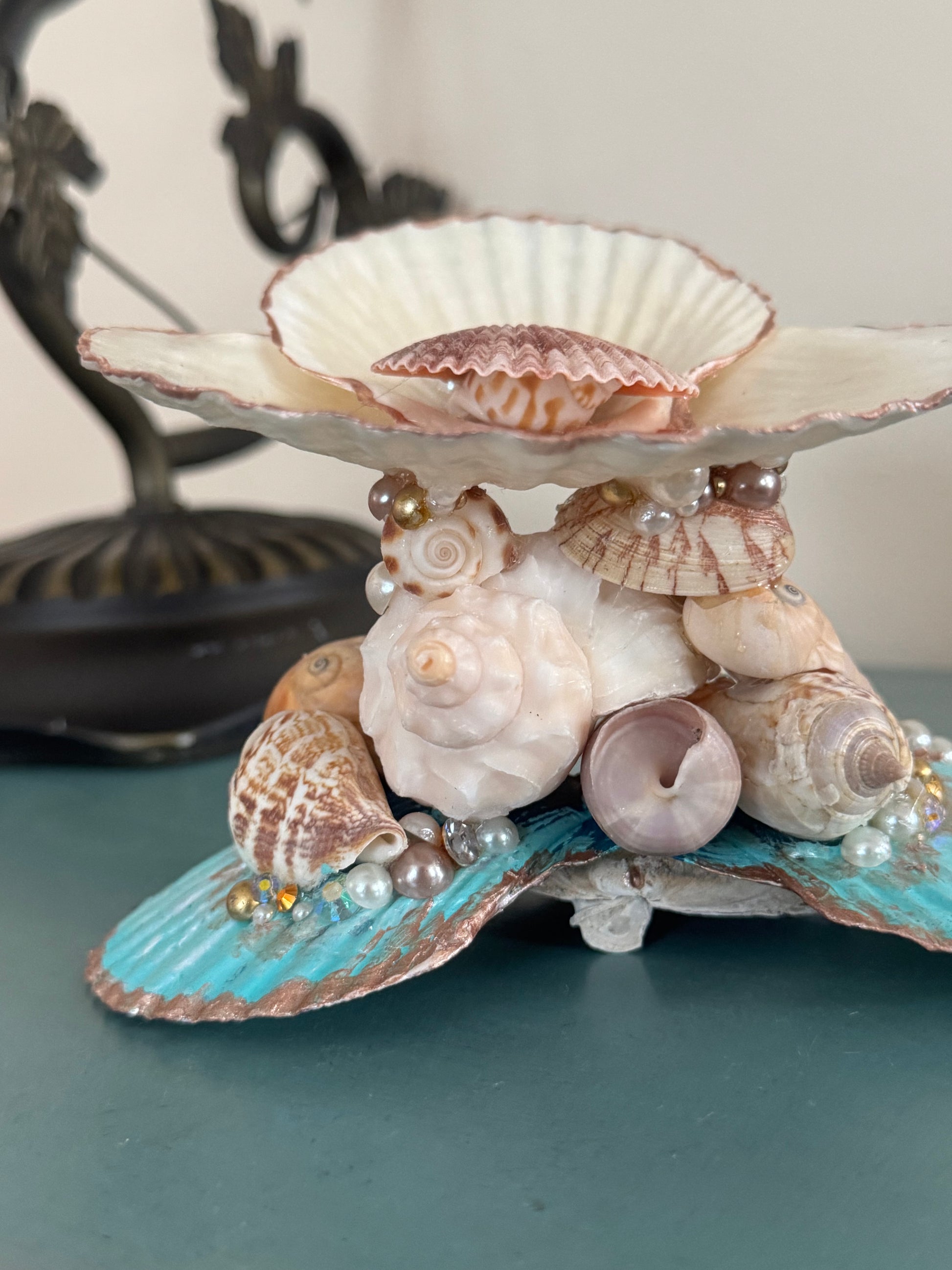 A decorative seashell and shell cluster stand with a scallop-shaped top tray, featuring various natural seashells, pearls, and small gold beads on a turquoise and white shell base, set against a dark teal surface.