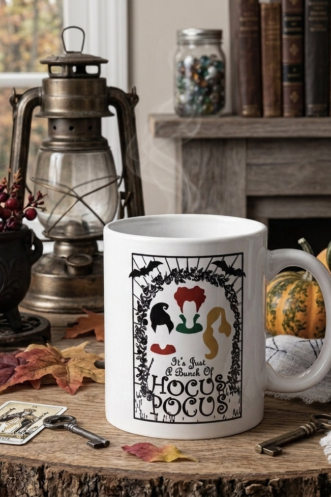 Sanderson sisters Halloween mug 'It's Gotta Be a Hocus Pocus Docus' sits on a rustic wooden table surrounded by autumn props including pumpkins, leaves, a lantern, and tarot cards.